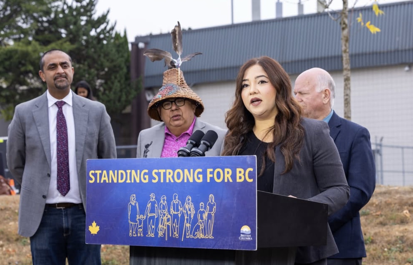Amna Shah speaking at an outdoor event beside a ‘Standing Strong for BC’ sign.